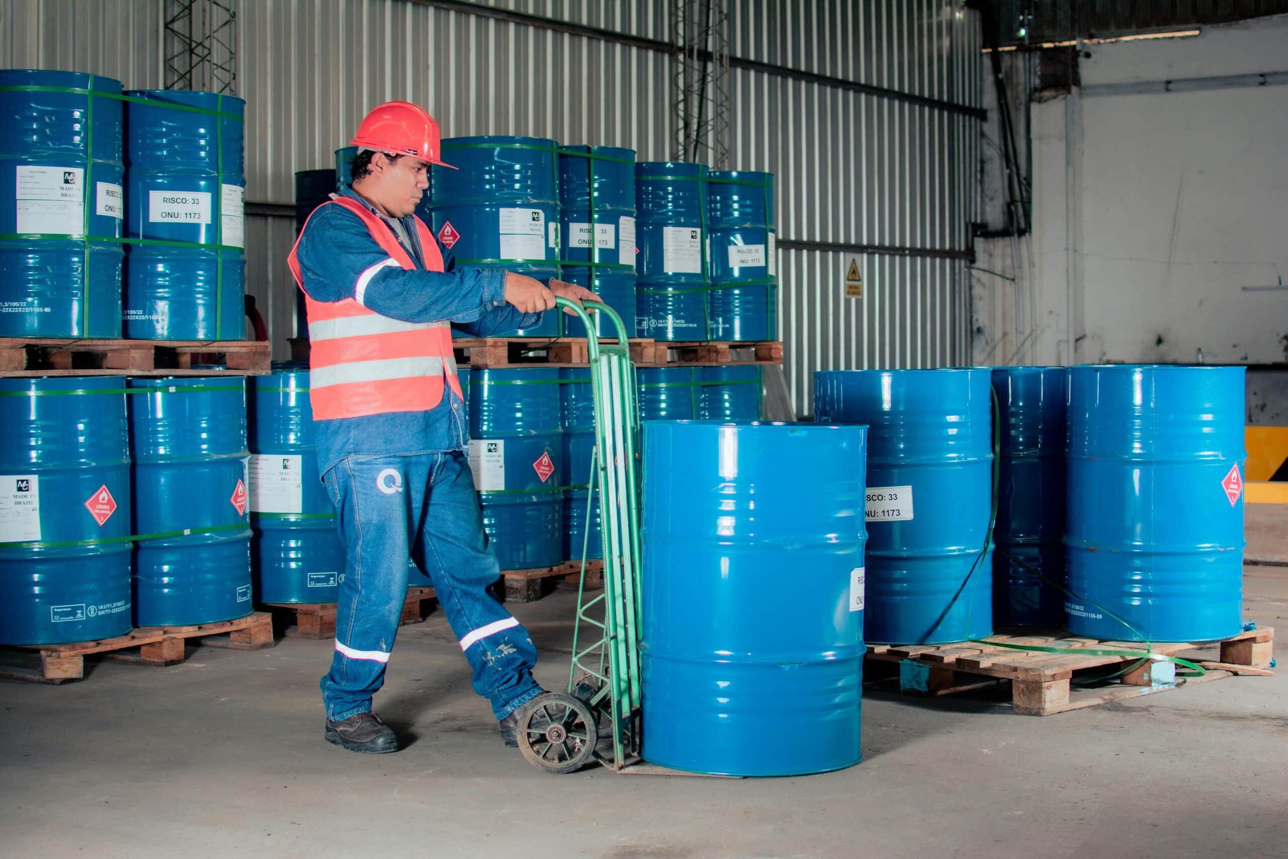 Ein Industriearbeiter, der mit einer Sackkarre blaue Fässer mit Chemikalien in einem Lagerraum transportiert.
